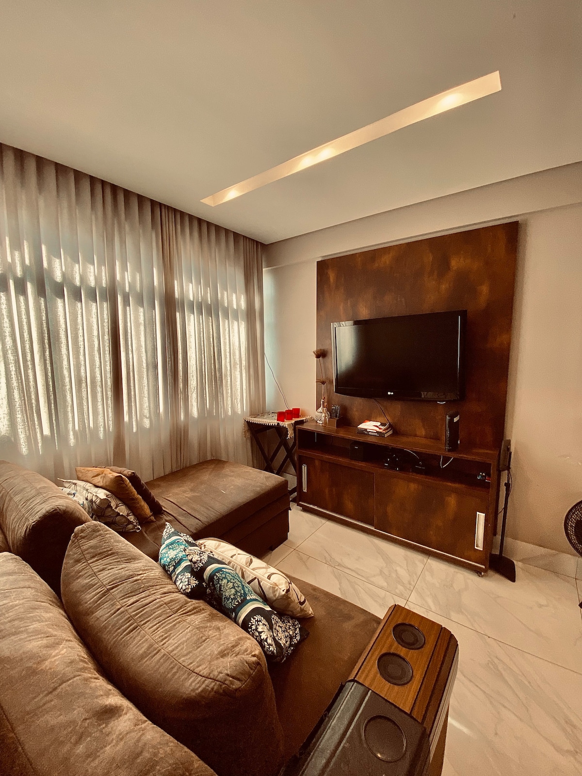 A cozy living area features a spacious sectional sofa adorned with decorative pillows. Natural light filters through sheer curtains, creating a soft ambiance. A dark wooden entertainment unit holds a flat-screen TV, while a small side table adds functionality to the space.