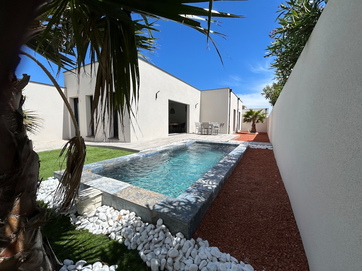 A serene outdoor area features a heated pool surrounded by smooth stones and lush green grass. The walls are accented with palm trees, and a dining table is visible on a patio in the background, providing a sense of open space and tranquility.