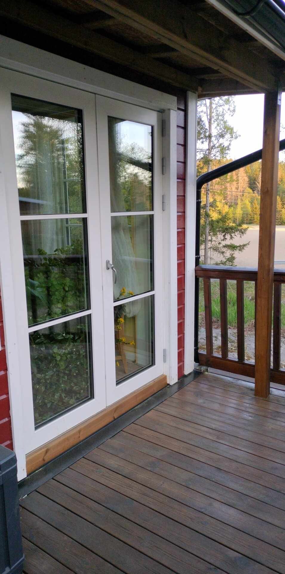 A deck area is presented with double glass doors that open into the cabin, framed by wooden railings. The warm wood tone of the flooring complements the red exterior of the cabin. Outside, greenery is visible in the background, enhancing the natural setting.