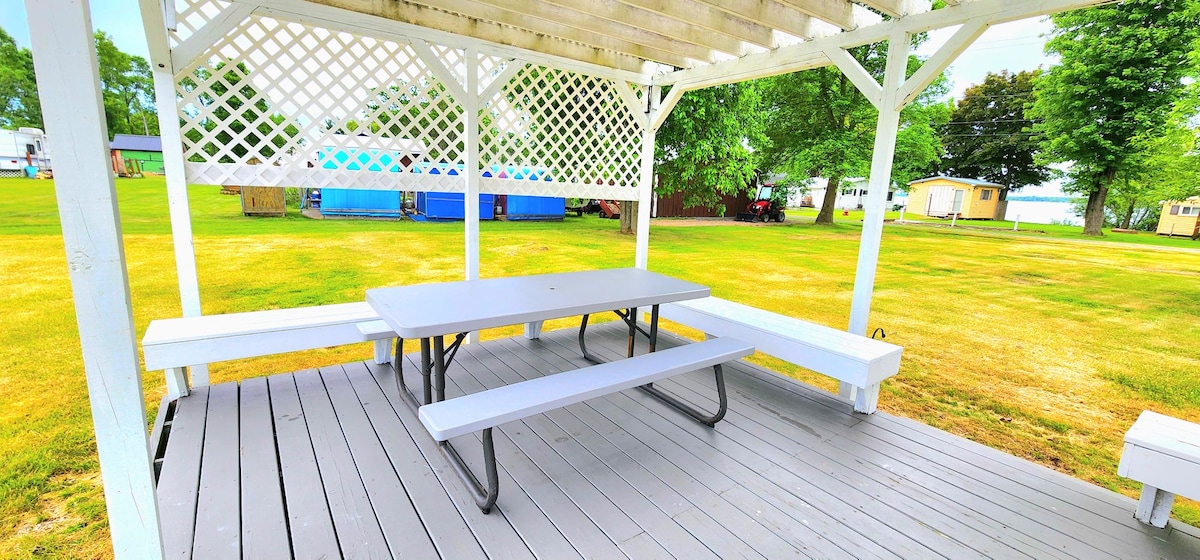 A covered picnic area is shown with a gray table and benches underneath a wooden lattice structure. The space offers a clear view of the lush green lawn surrounding it, providing a serene spot for outdoor meals or gatherings.