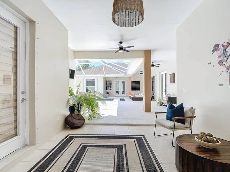An inviting entryway is framed by a light-colored wall and topped with a woven pendant lamp. A comfortable chair sits beside a round wooden table. Natural light illuminates the space, highlighting the outdoor area visible through large sliding doors and potted greenery.
