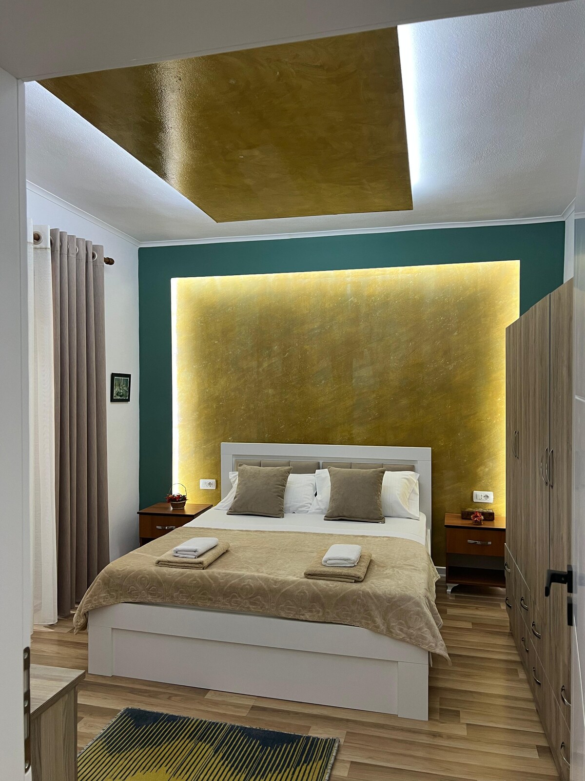 A serene bedroom features a spacious bed with soft cream linens and accent pillows. A textured gold wall is lit subtly, enhancing the ambiance. Light wood nightstands are positioned on either side of the bed, while soft drapes frame a window, creating a calming atmosphere.