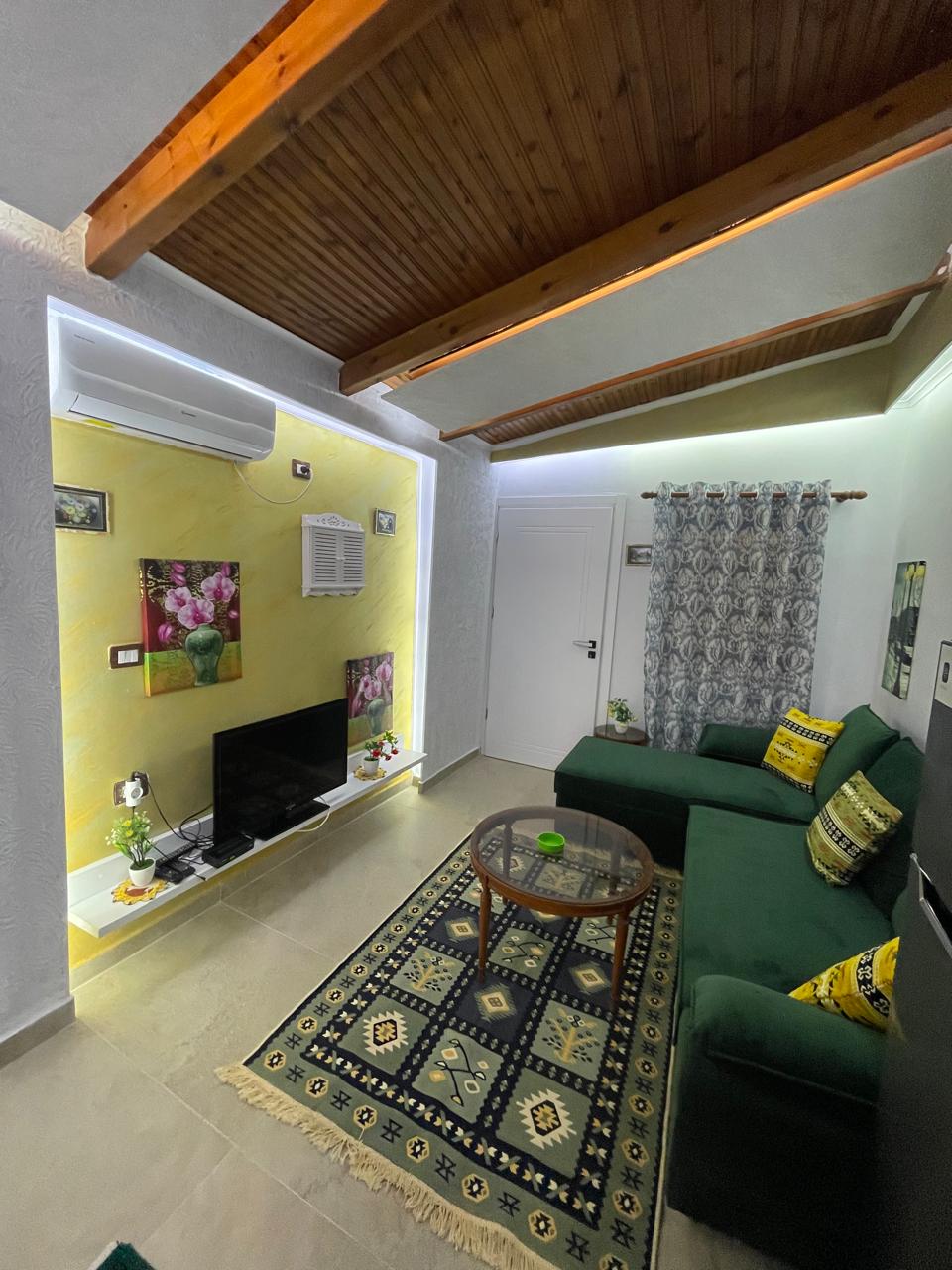 A living area is presented with a green sofa and a glass coffee table on a patterned rug. The walls feature floral artwork, and a television is positioned opposite the seating. Bright lighting highlights the wooden ceiling and adds warmth to the space.
