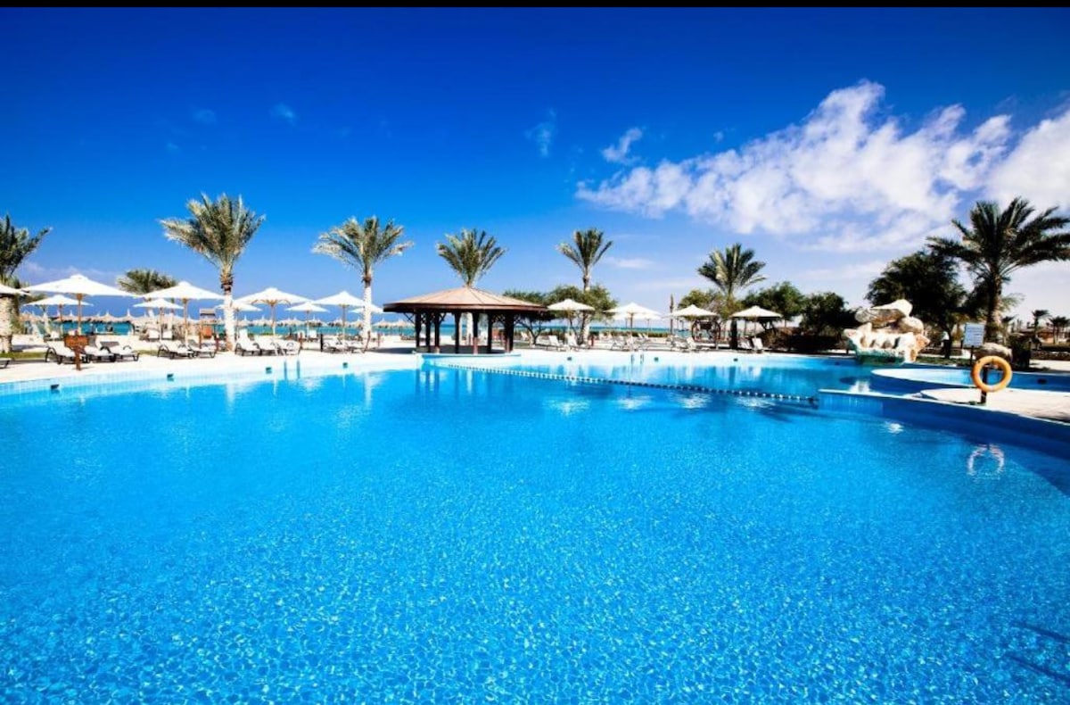 A large swimming pool is surrounded by swaying palm trees and sun loungers. A shaded structure sits nearby, providing a relaxing spot for guests. Clear blue skies reflect off the water's surface, contributing to a serene outdoor environment.