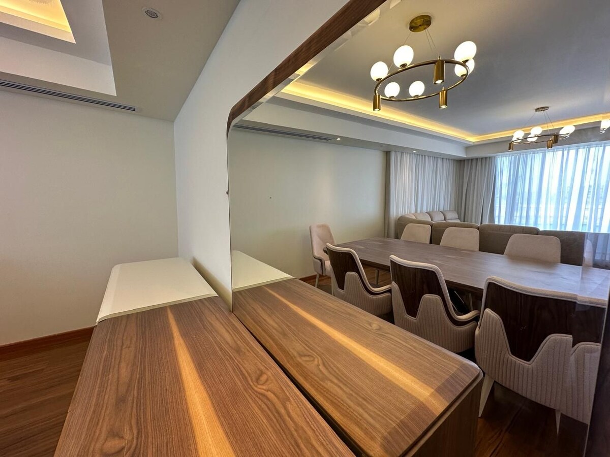A dining area is showcased, featuring a large table surrounded by well-upholstered chairs. A modern buffet unit is located nearby, and soft lighting from an overhead fixture enhances the space's ambiance. Large windows allow natural light to brighten the room.