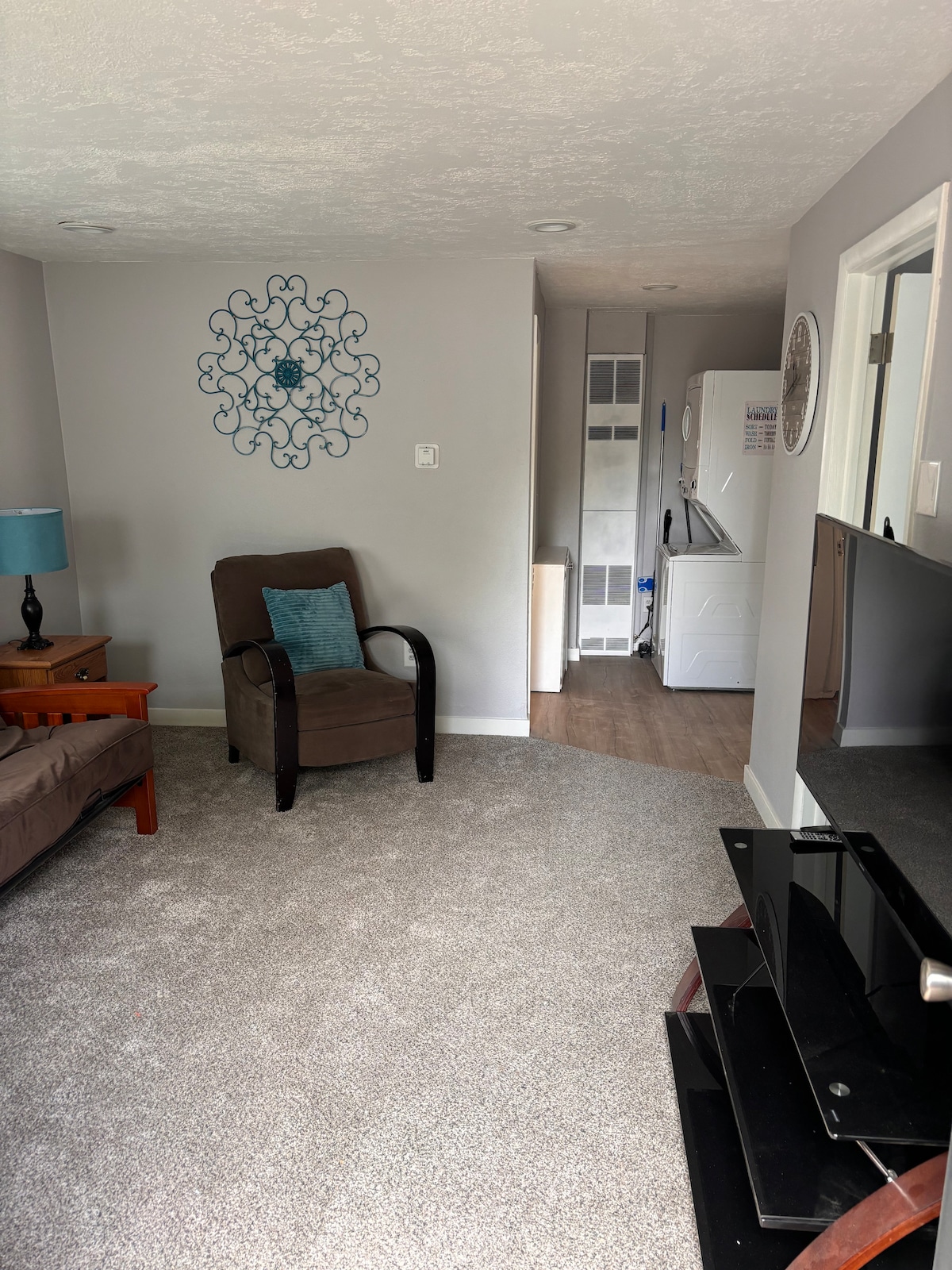 A cozy living area features a comfortable chair and a futon, accompanied by a small side table with a blue lamp. A decorative wall piece is mounted above the seating. In the background, a doorway leads to a laundry area complete with a washer and dryer.