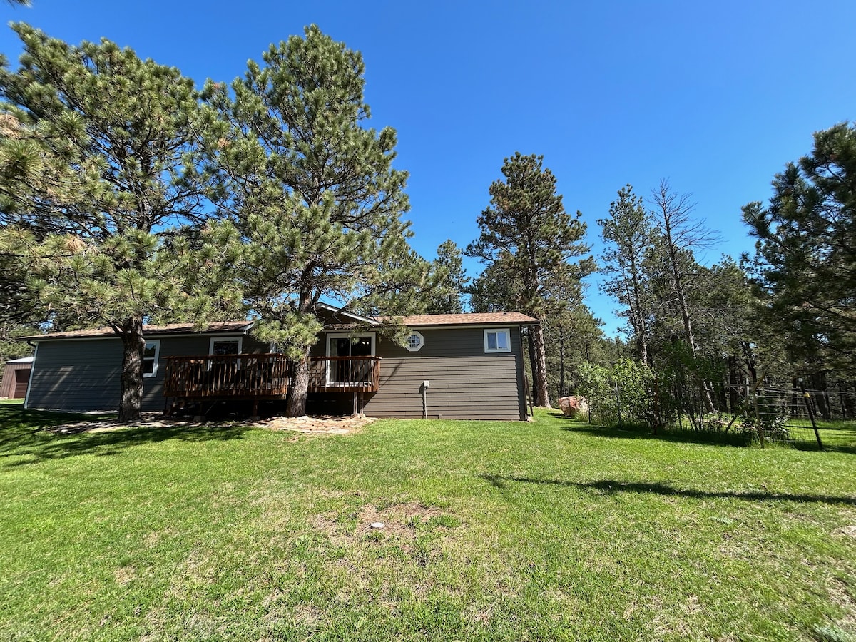The exterior of a single-story home is framed by tall pine trees, with a well-maintained grassy area in the foreground. A wooden deck extends from the house, inviting outdoor relaxation, while the clear blue sky creates a bright backdrop.