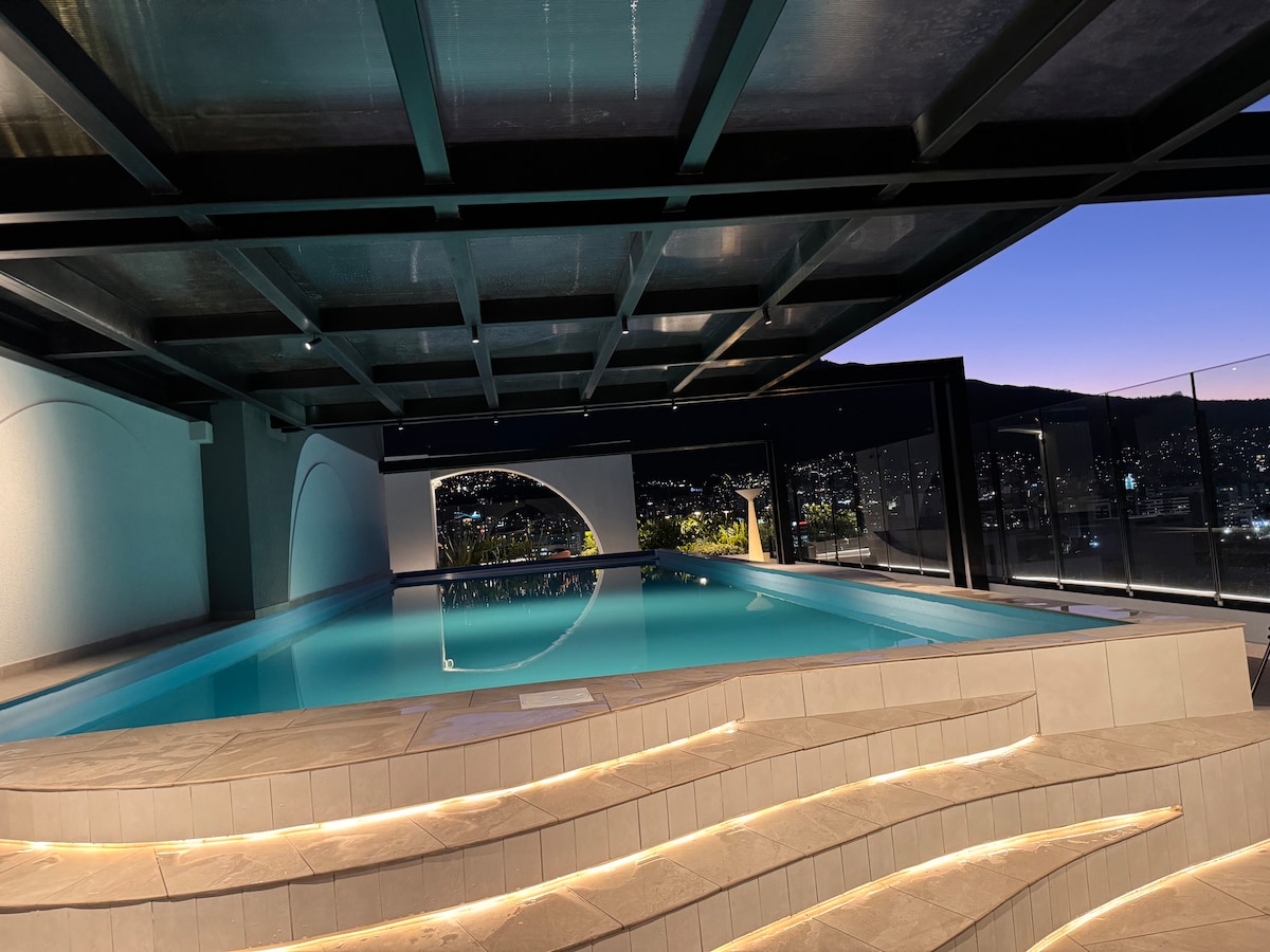 An indoor swimming pool is depicted, featuring a sleek design with smooth, illuminated stairs leading into the water. Large glass panels showcase a nighttime view of the city skyline, creating a serene and modern environment for guests.