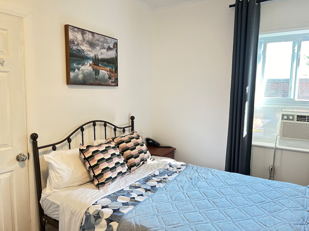 A bedroom features a double-sized bed with patterned pillows and a soft blue quilt. A piece of landscape artwork decorates the wall above the bed. Natural light enters through a window to the right, beside an air conditioning unit. A dark curtain frames the window.