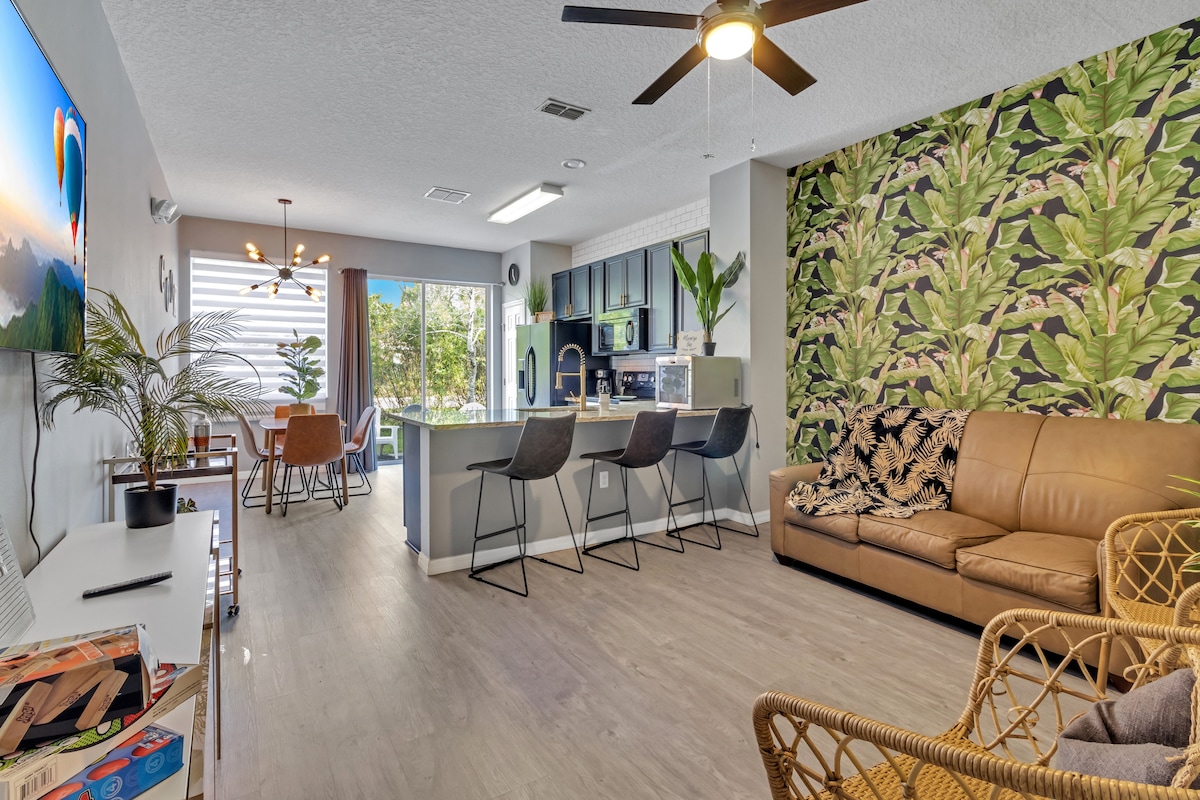 A spacious living area features a brown leather sofa and intricate rattan chairs, creating comfortable seating arrangements. A vibrant plant-themed wallpaper adds a refreshing touch. The adjoining dining space is visible, along with modern kitchen appliances that blend seamlessly with the open layout.