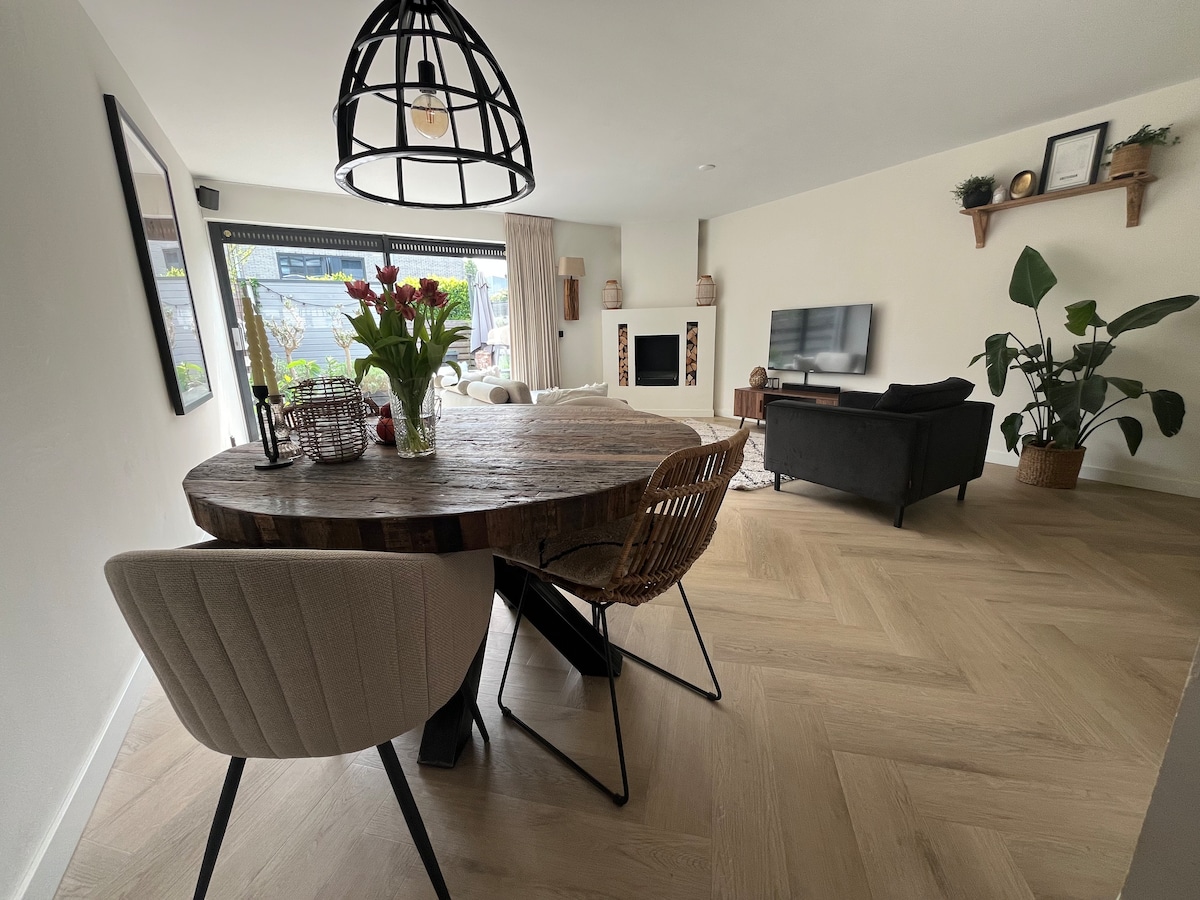 A dining area is presented with a rustic wooden table surrounded by elegant chairs. Natural light enters through large windows, enhancing the space. A TV unit and a comfortable sofa are visible, complemented by indoor plants that add a touch of greenery.