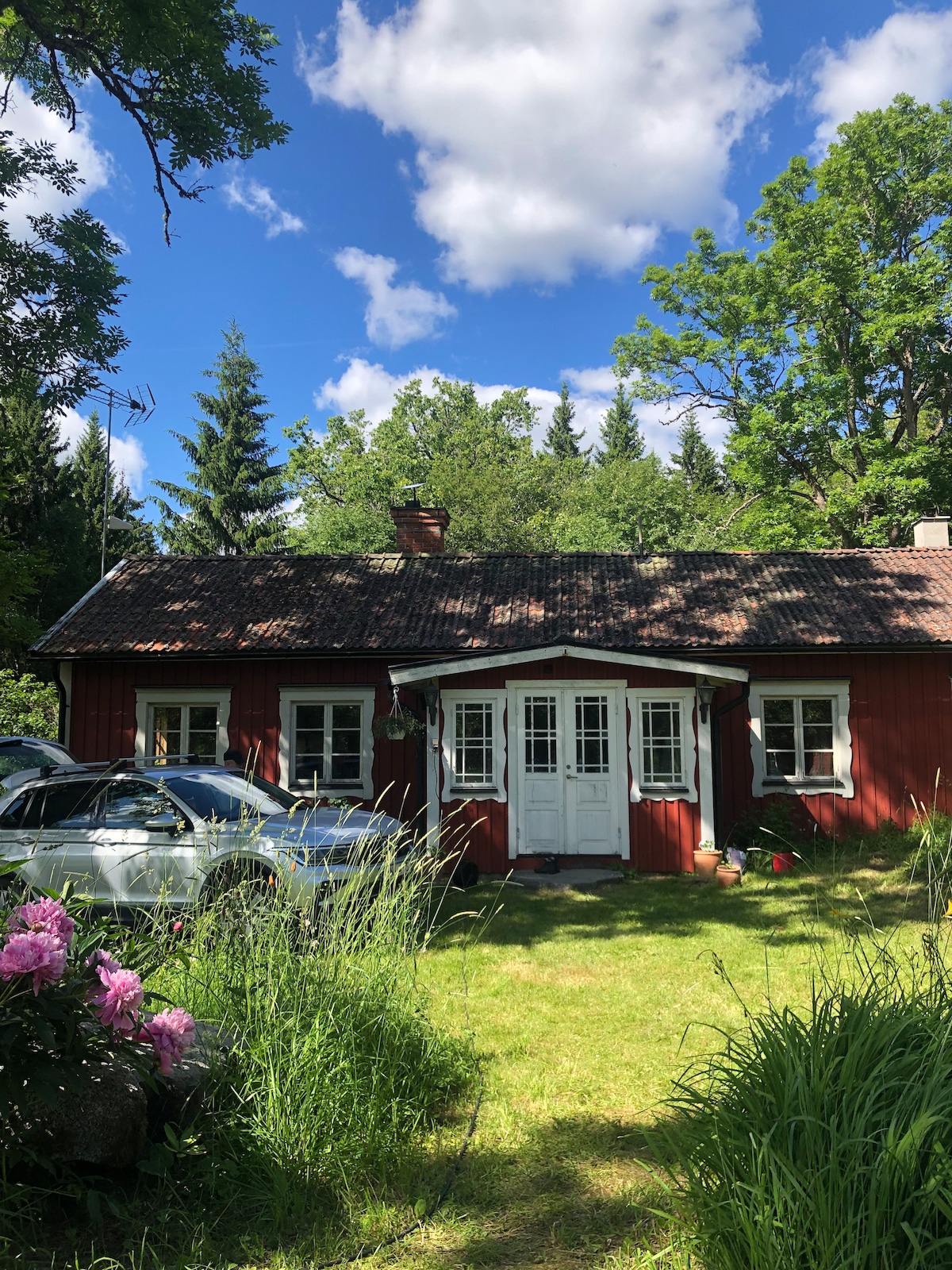A charming red house is set amidst lush greenery, surrounded by trees and colorful flowers. The property features a well-maintained lawn, with a clear blue sky overhead and scattered clouds. Two cars are parked nearby, enhancing the cozy rural atmosphere.