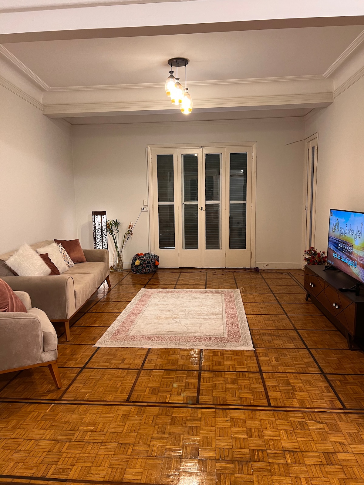 A living area features a soft, neutral-toned sofa with decorative cushions, positioned across from a wooden television stand. A rectangular rug is centered on the polished wooden floor, while natural light enters through glass doors that lead to a balcony. A floor lamp adds subtle illumination.