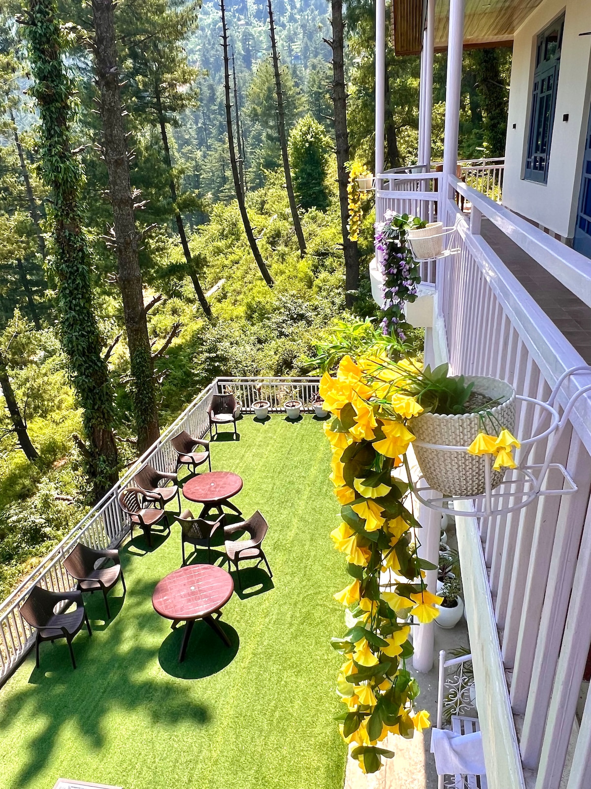 A balcony overlooks a lush green landscape, featuring multiple round tables surrounded by chairs. Flower pots hang from the railing, displaying bright yellow blooms. The area is covered with green turf, enhancing the outdoor relaxation space.