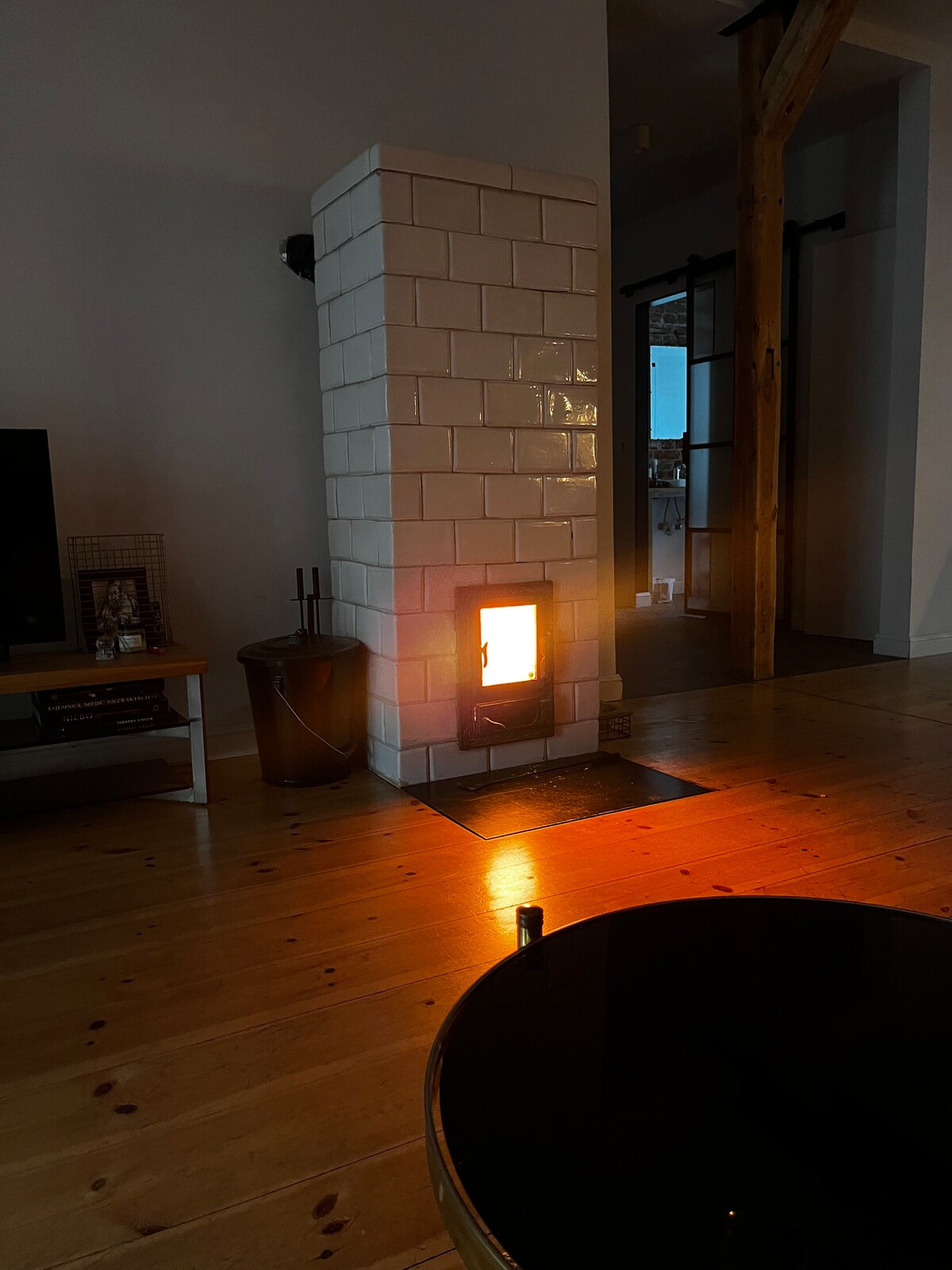 A well-lit living area features a modern fireplace, accentuating the warm glow of the flames. Light reflects off the polished wooden floor, while a dark coffee table sits nearby. Visible in the background is a doorway leading to additional rooms.