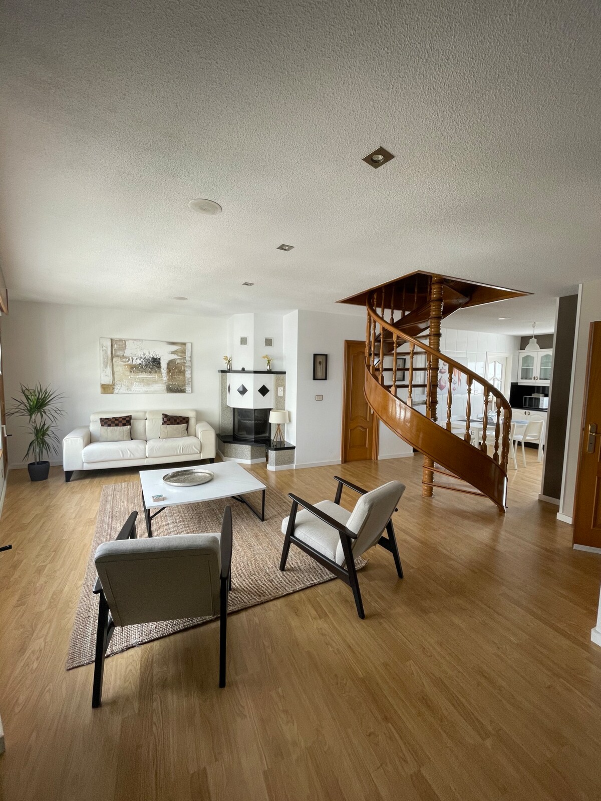 A spacious living area features a white sofa and two armchairs surrounding a light-colored coffee table. A decorative painting is displayed on the wall, and a potted plant adds a touch of greenery. A wooden spiral staircase leads to the upper floor, enhancing the open layout.