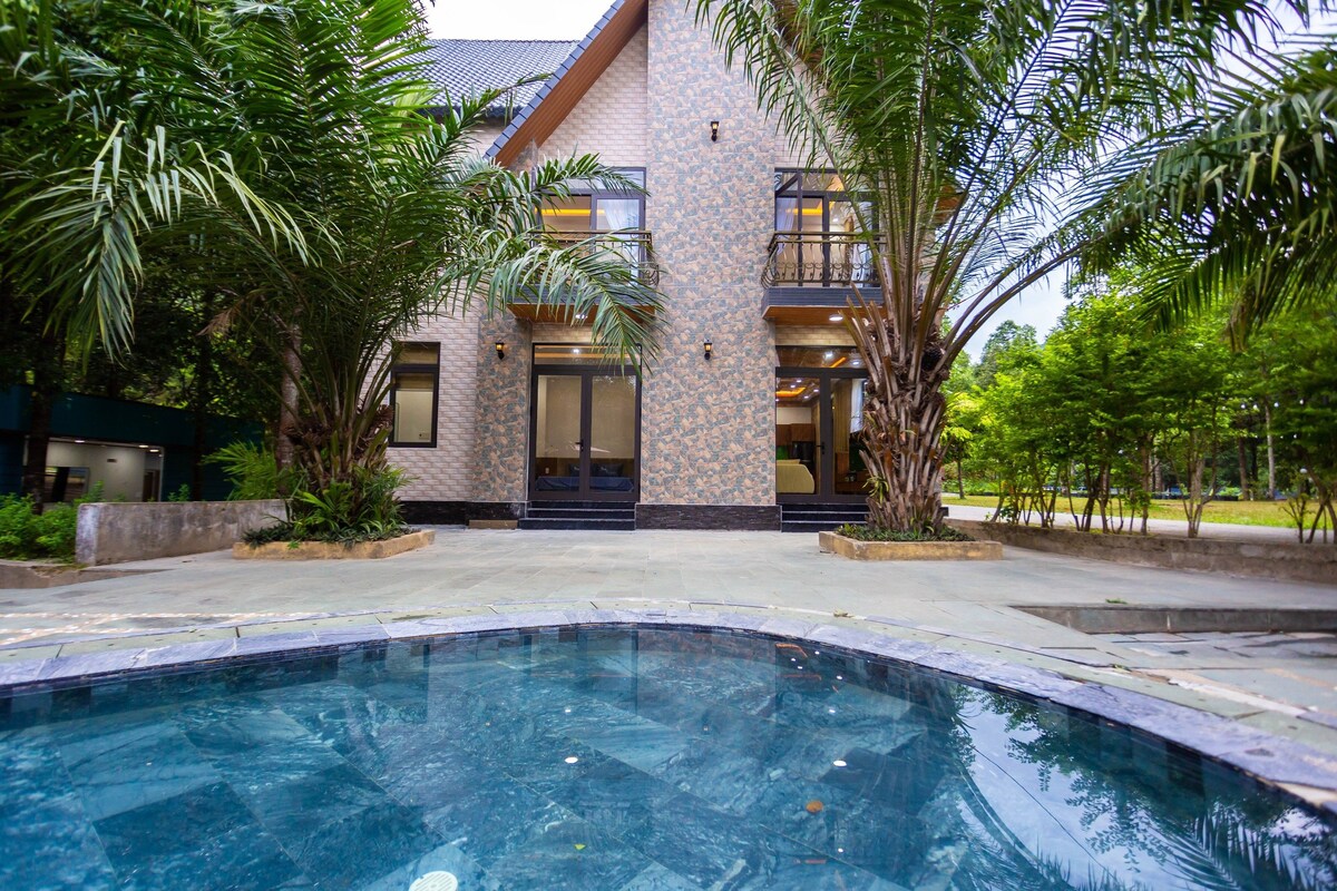 The villa is surrounded by lush palm trees, with two floors visible through large glass doors. A private swimming pool reflects the exterior's colors. The outdoor area is paved with stones and includes a spacious layout, enhancing the connection to the natural surroundings.