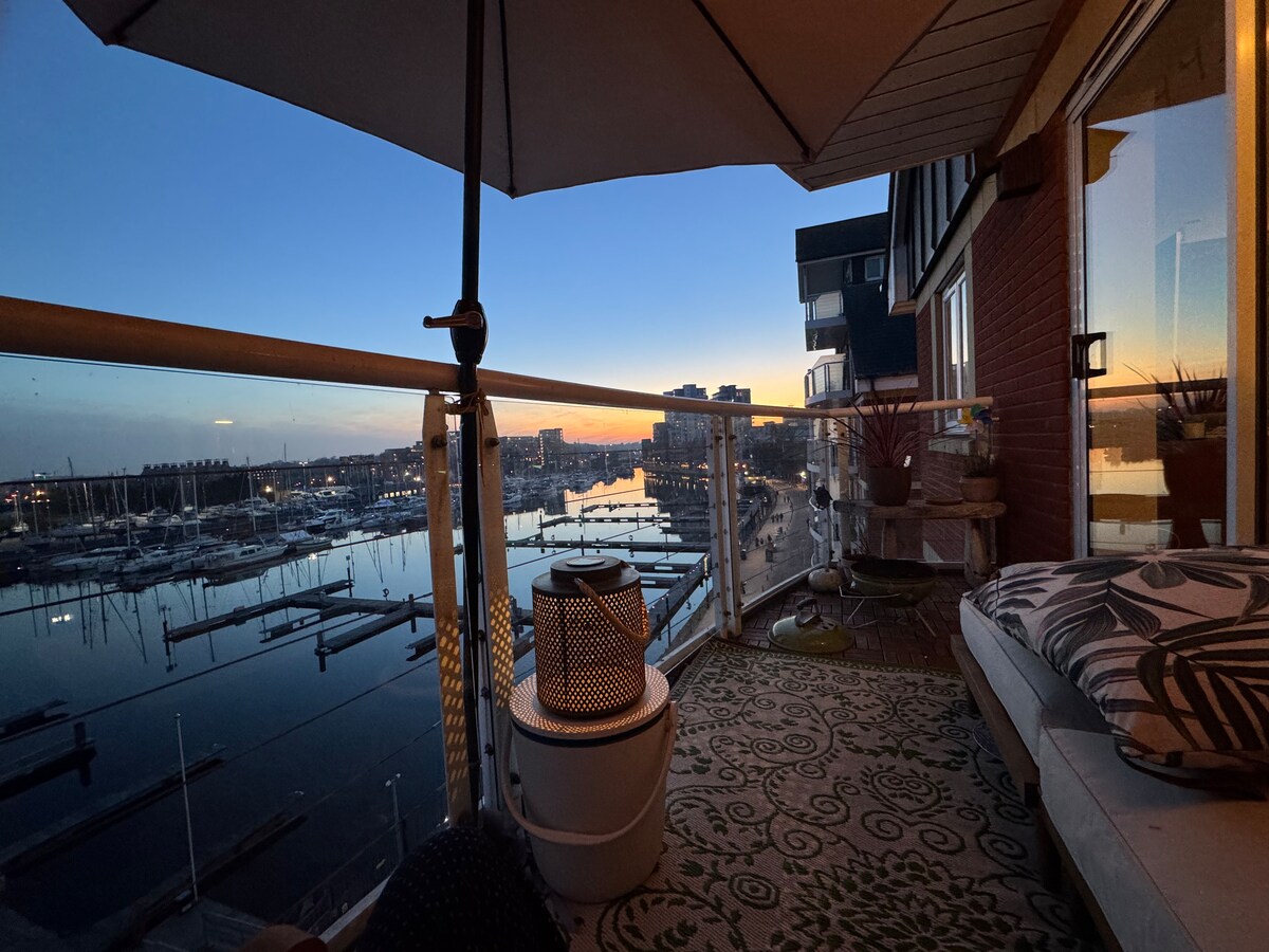 A balcony overlooking the tranquil marina features a cozy seating area, adorned with cushions and patterned textiles. The scene is framed by an umbrella, with water reflections from the sunset creating a serene atmosphere. The distant cityscape adds to the scenic view.