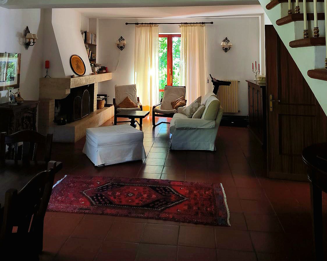 A spacious living area features a warm-toned tile floor and large windows that allow natural light to fill the room. Comfortable seating arrangements are highlighted by two armchairs and a sofa. A traditional area rug adds color, and a fireplace is positioned near the wall.
