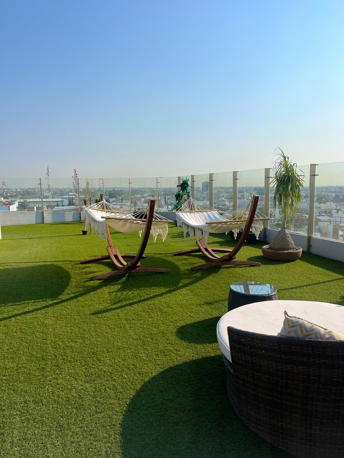 A rooftop area features two hammocks suspended on wooden stands, surrounded by a synthetic grass surface. Potted plants add a touch of greenery, and a circular seating arrangement is visible in the foreground, offering a serene space with expansive city views.