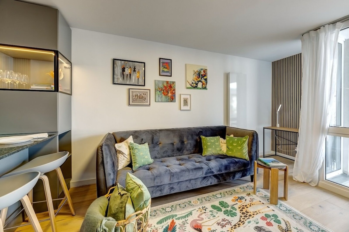 A living area is presented with a plush sofa adorned with decorative cushions, complemented by a light-colored area rug. Bright artwork is displayed on the walls, and a small side table is positioned next to a floor lamp. Natural light filters through large windows, enhancing the space.