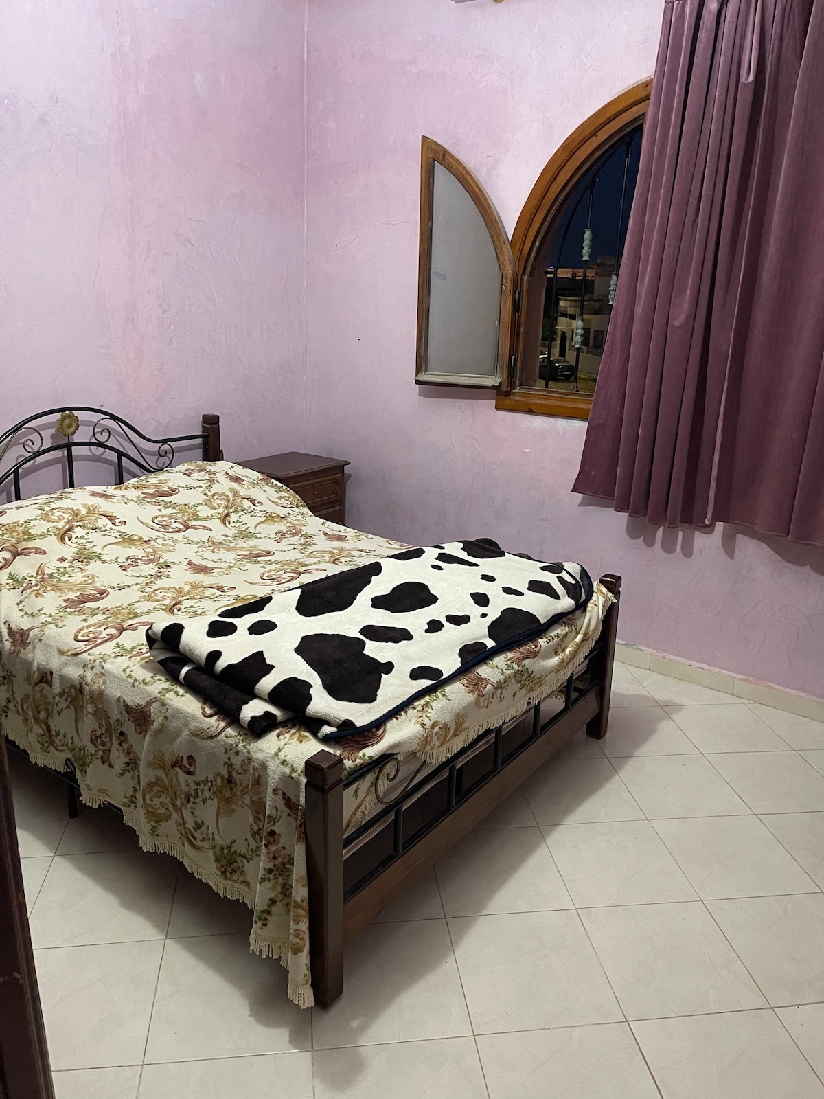 A softly lit bedroom features a spacious bed adorned with a floral coverlet and a patterned blanket. The room is painted in a light pink hue, with a large window framed by dark curtains and a nightstand placed beside the bed.