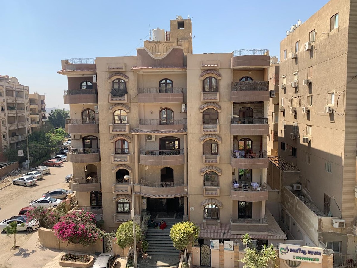 The exterior of a four-story residential building is shown, characterized by arched balconies and decorative features. Surrounding greenery includes small trees and flowering plants, enhancing the entrance area. A parking lot is visible in the foreground, providing spaces for vehicles.