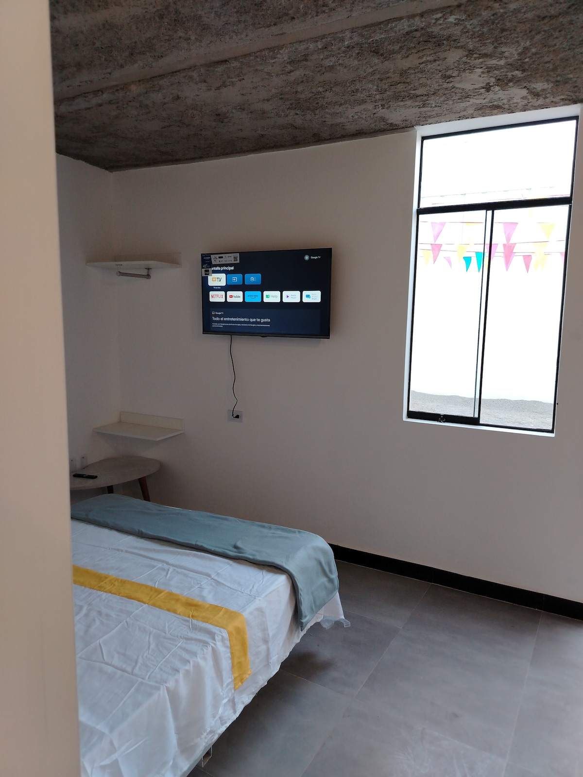 A bright sleeping area features a spacious bed dressed in white linen with a yellow accent. A wall-mounted television is positioned across from the bed. A narrow desk is situated nearby under an open window adorned with colorful banners.