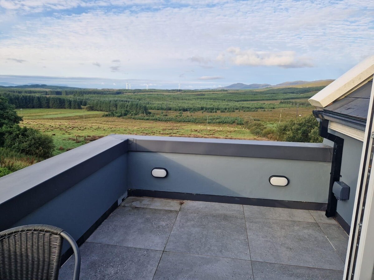 A spacious outdoor terrace offers unobstructed views of the lush, rolling countryside, punctuated by distant hills. The stone floor creates a modern touch, and a small round table with a chair is positioned in a corner, inviting relaxation amidst the serene landscape.