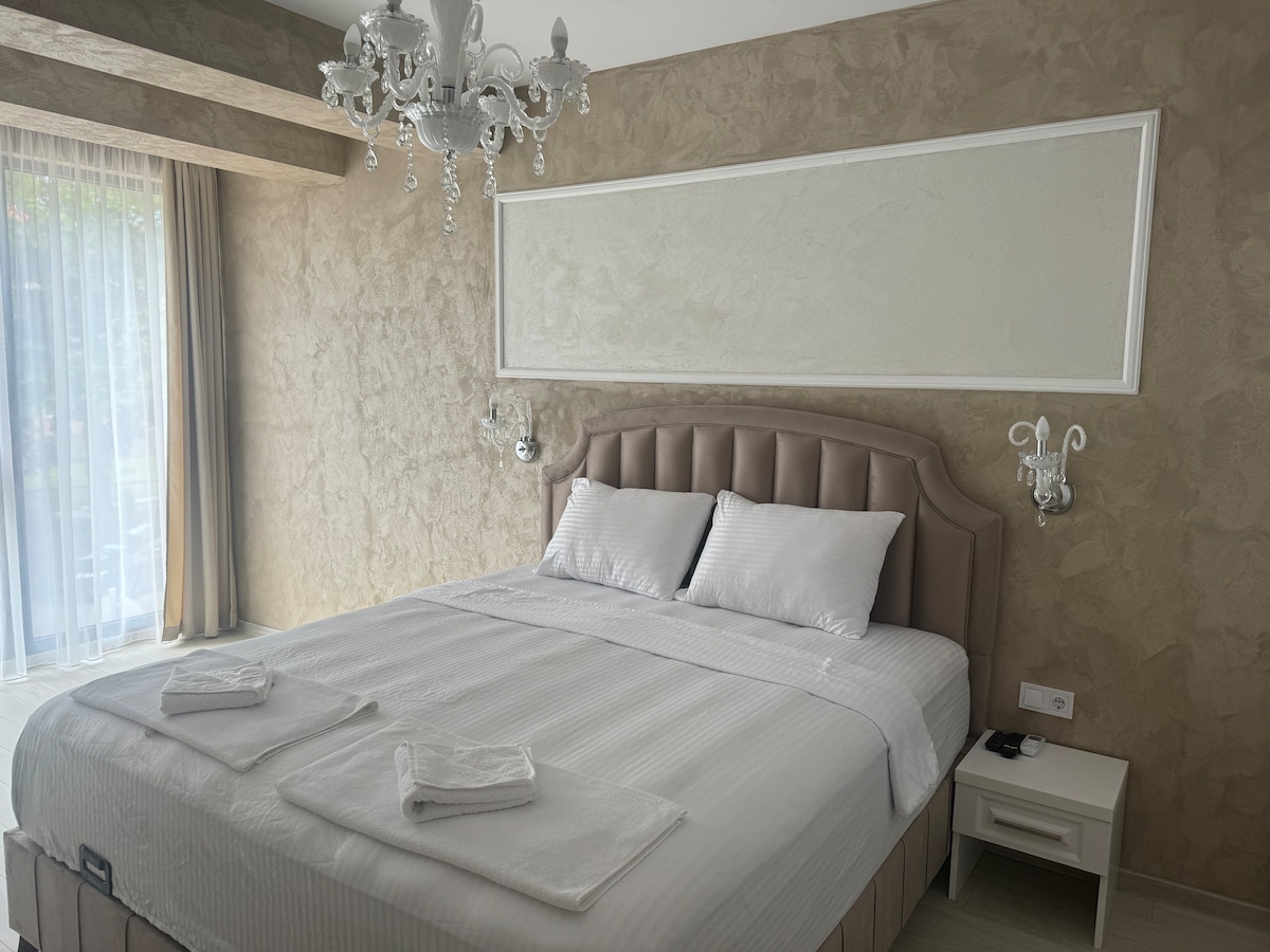 A spacious bed is centered in the room, complemented by soft, neutral-toned linens. Two bedside lamps are positioned symmetrically on either side. Natural light filters through sheer curtains, highlighting the textured wall and elegant chandelier above.