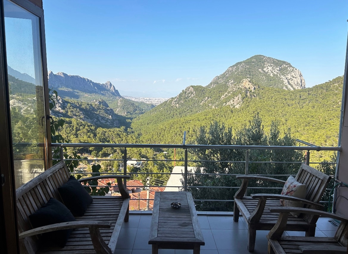 A spacious balcony features wooden seating, surrounded by scenic views of lush green mountains and valleys. The glass doors provide a clear connection to the outdoors, with a distant city skyline visible under a bright blue sky.