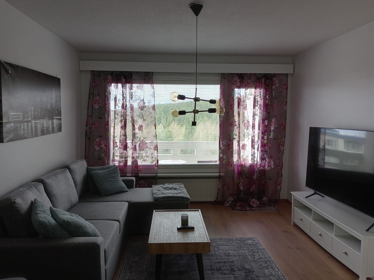 A modern living area is presented, featuring a cozy grey sofa adorned with decorative cushions. Large windows, covered with floral curtains, provide ample natural light and a view of the surroundings. A simple coffee table and a white entertainment unit complement the space.