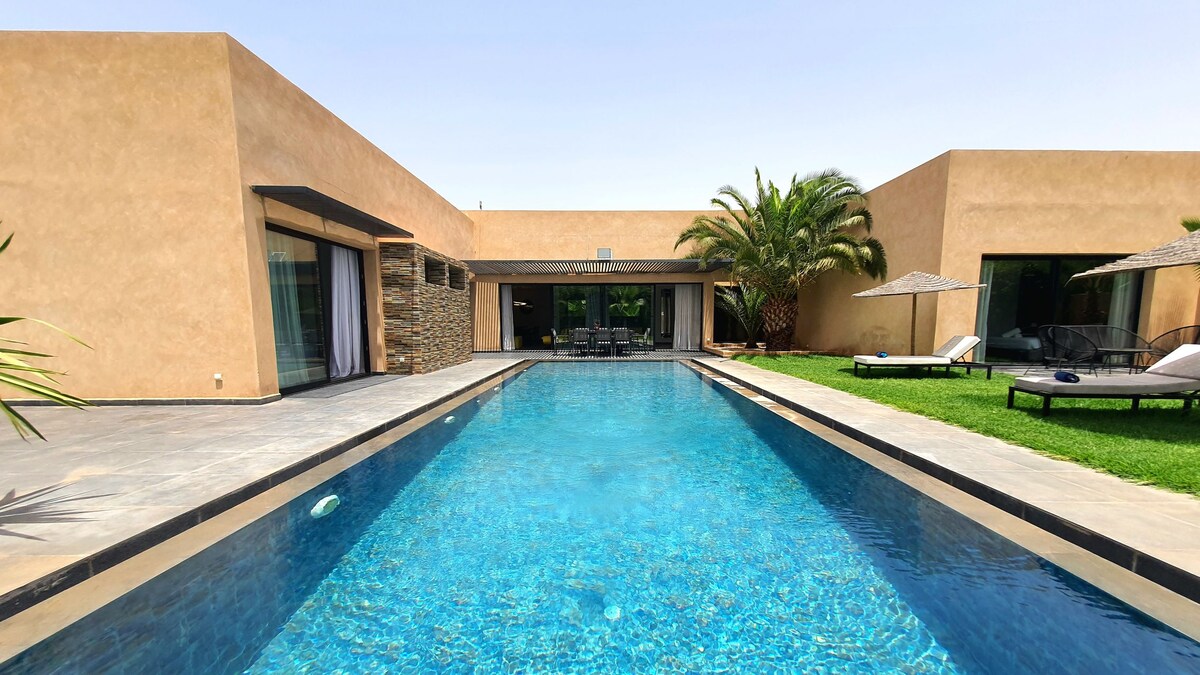 An expansive swimming pool reflects the clear sky, surrounded by well-maintained grass. The villa's elegant facade features large windows that offer views of the outdoor space. Chaise lounges are positioned nearby, with a shaded area provided by a parasol for relaxation.