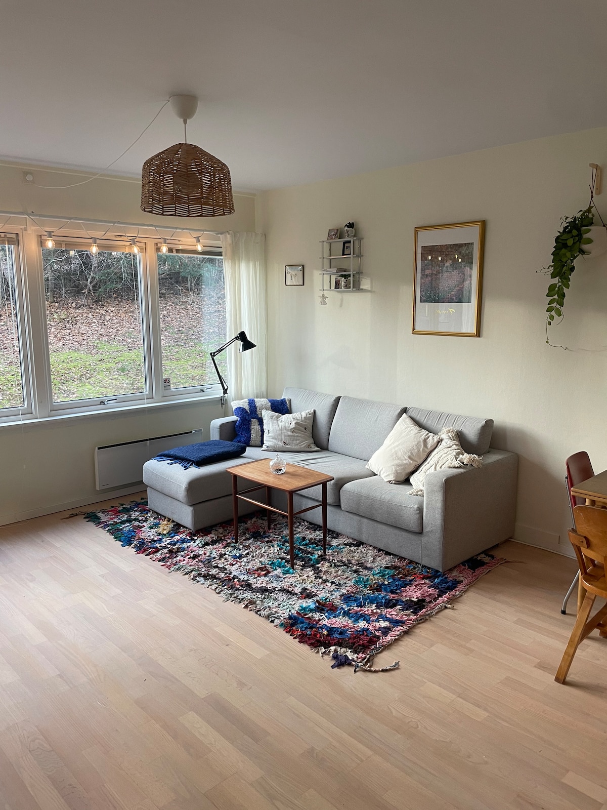 A living area is displayed with a light-colored sofa adorned with decorative pillows. A small wooden table is positioned in front of the sofa, set on a colorful rug. Natural light enters through large windows, illuminating the space and showcasing a greenery accent.