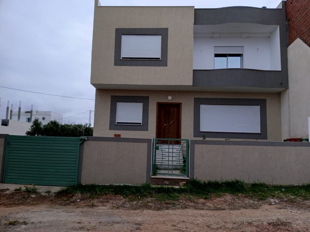The exterior of a modern duplex house is presented, featuring a clean façade with grey and white accents. Large windows are seen, complemented by a gated entrance. A small area of landscaped earth borders the front of the house, contributing to a neat outdoor appearance.