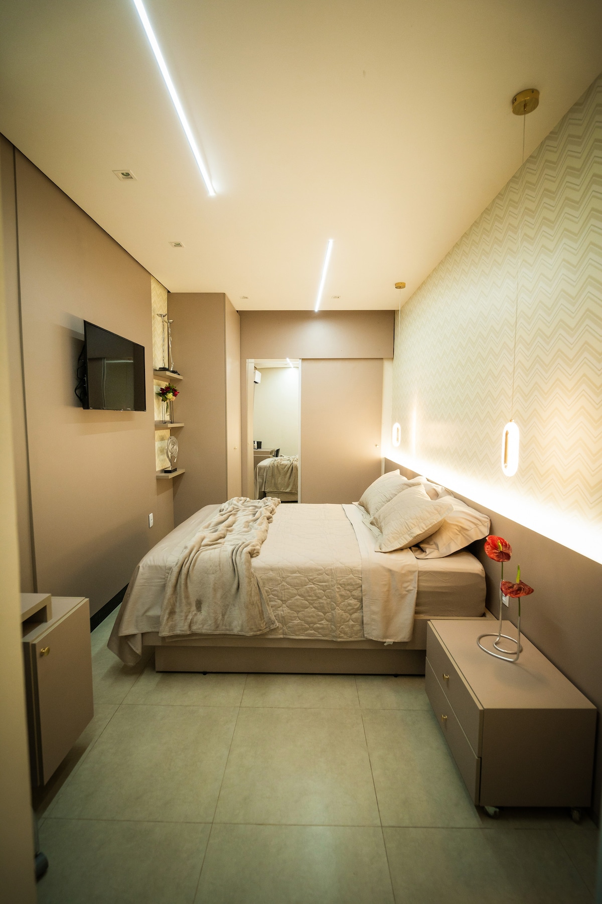 A well-appointed bedroom features a spacious bed adorned with soft linens. Wall sconces provide gentle lighting, and a television is mounted on the wall opposite the bed. A bedside table with a small vase and a blooming flower adds a touch of warmth to the space.