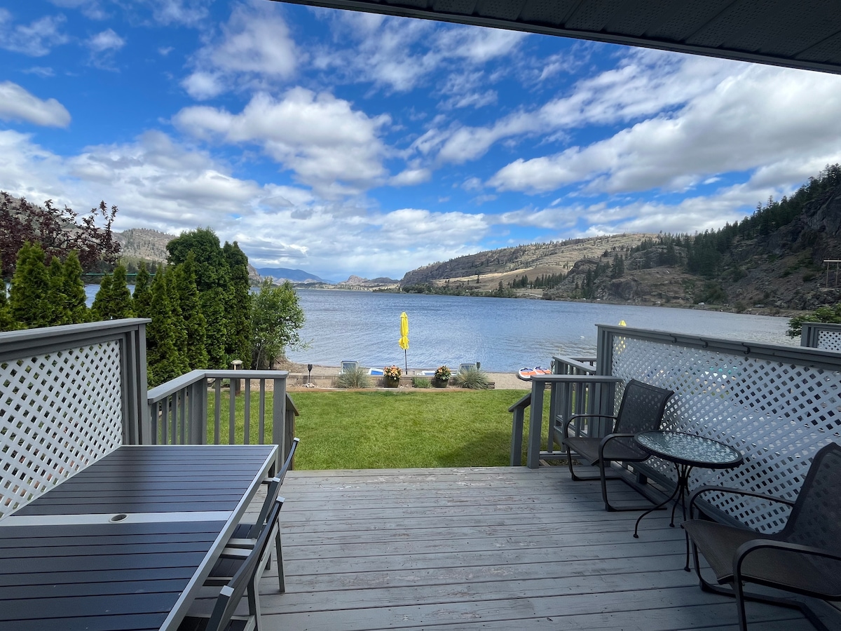 A private deck overlooks Vaseux Lake, framed by lush greenery and sandy beachfront. A yellow umbrella stands on the shore, accompanied by accessible watercraft. Comfortable seating arrangements are positioned on the deck, inviting relaxation while enjoying the scenic views and open sky.