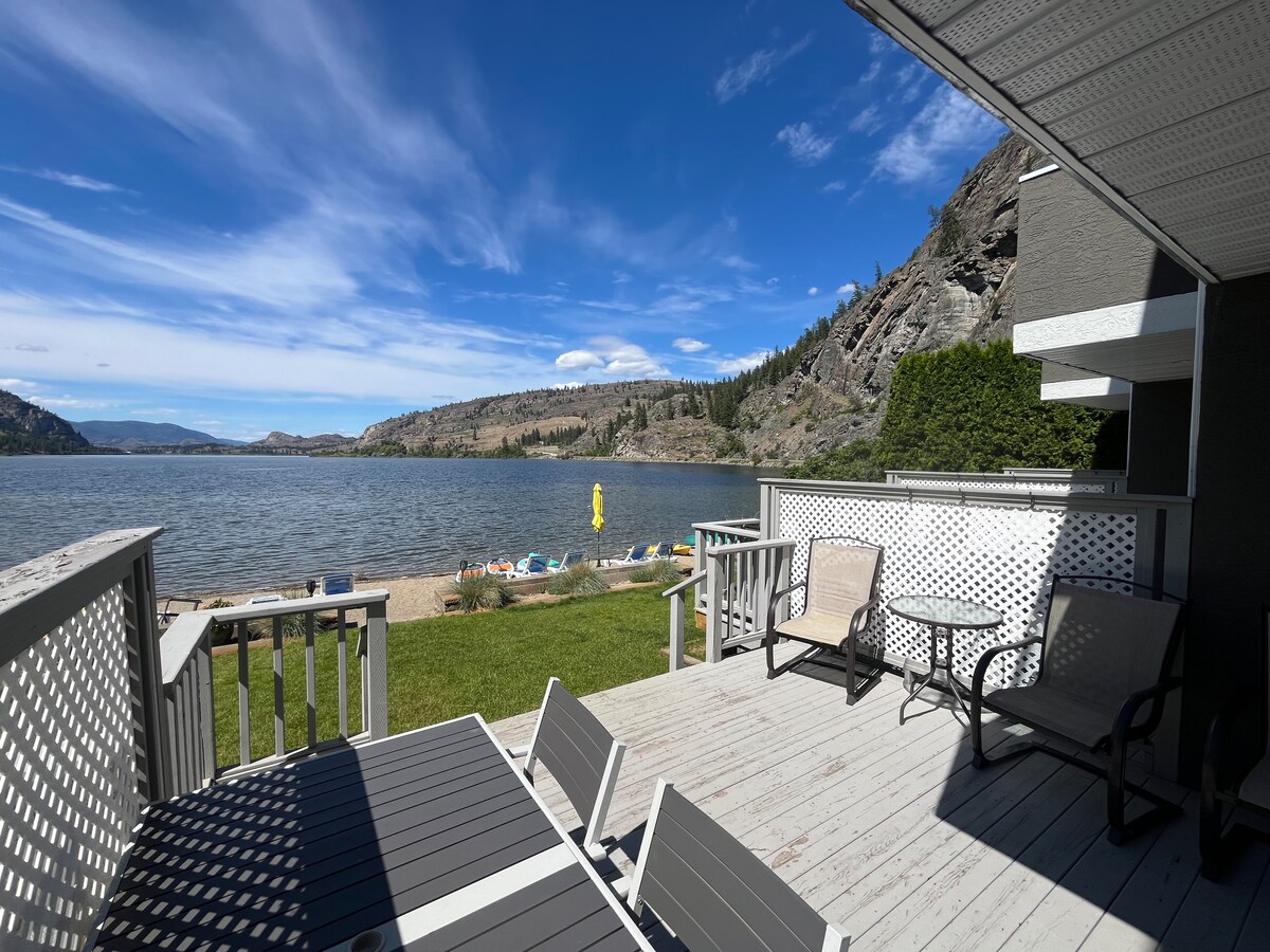 A private deck overlooks Vaseux Lake, featuring seating arrangements including lounge chairs. The sandy beachfront is visible, with kayaks and paddle boards arranged along the shore. Scenic hills and blue skies create a serene backdrop, enhancing the outdoor space for relaxation.
