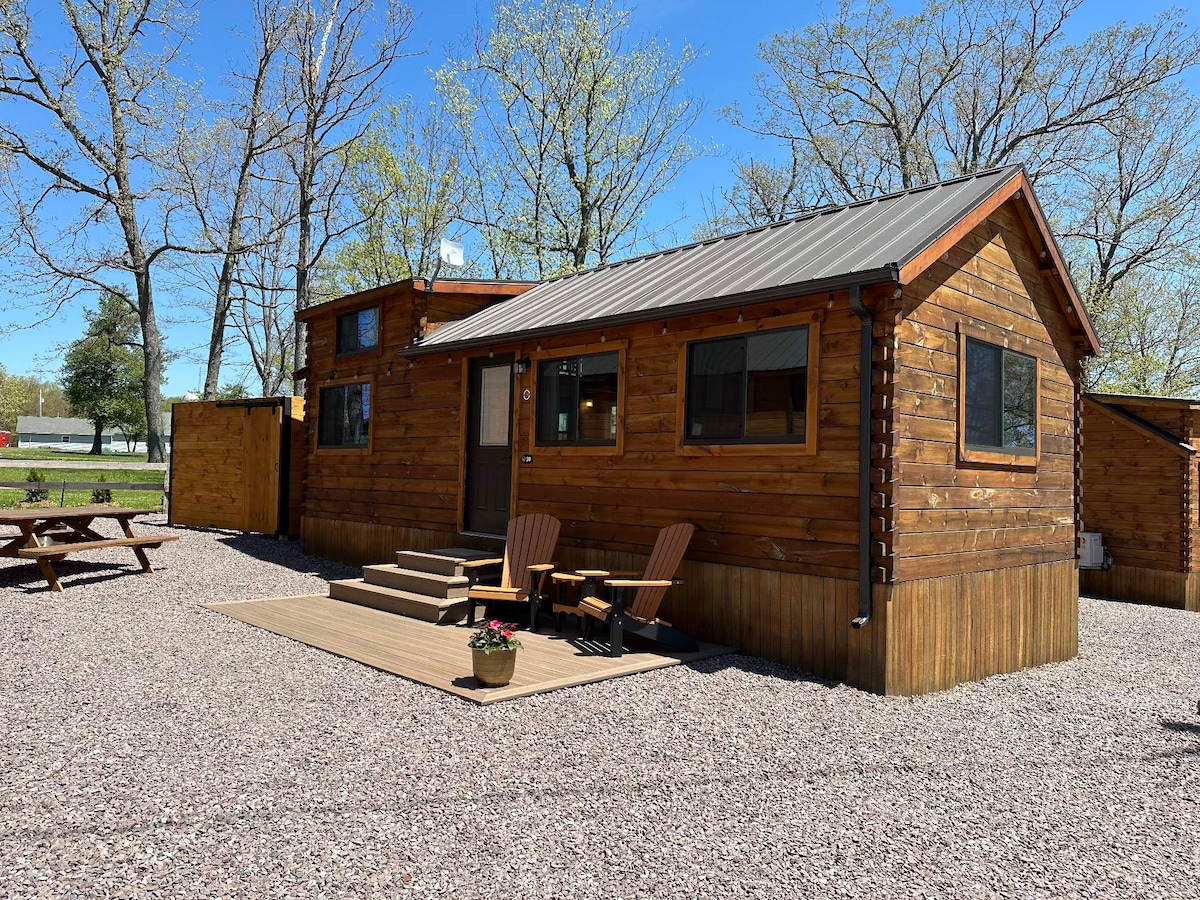 A log cabin is set against a backdrop of clear blue skies and trees. The structure features wood siding and a metal roof. A small deck with two chairs and a potted plant welcomes guests, while gravel landscaping surrounds the cabin.