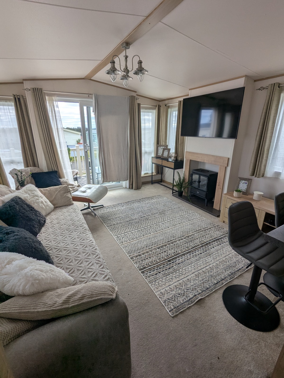 The living area is furnished with a cozy sectional sofa adorned with pillows, complemented by a patterned rug. A mounted television is positioned above a fireplace, while large windows allow natural light to fill the space. A dining chair is placed at the small desk.