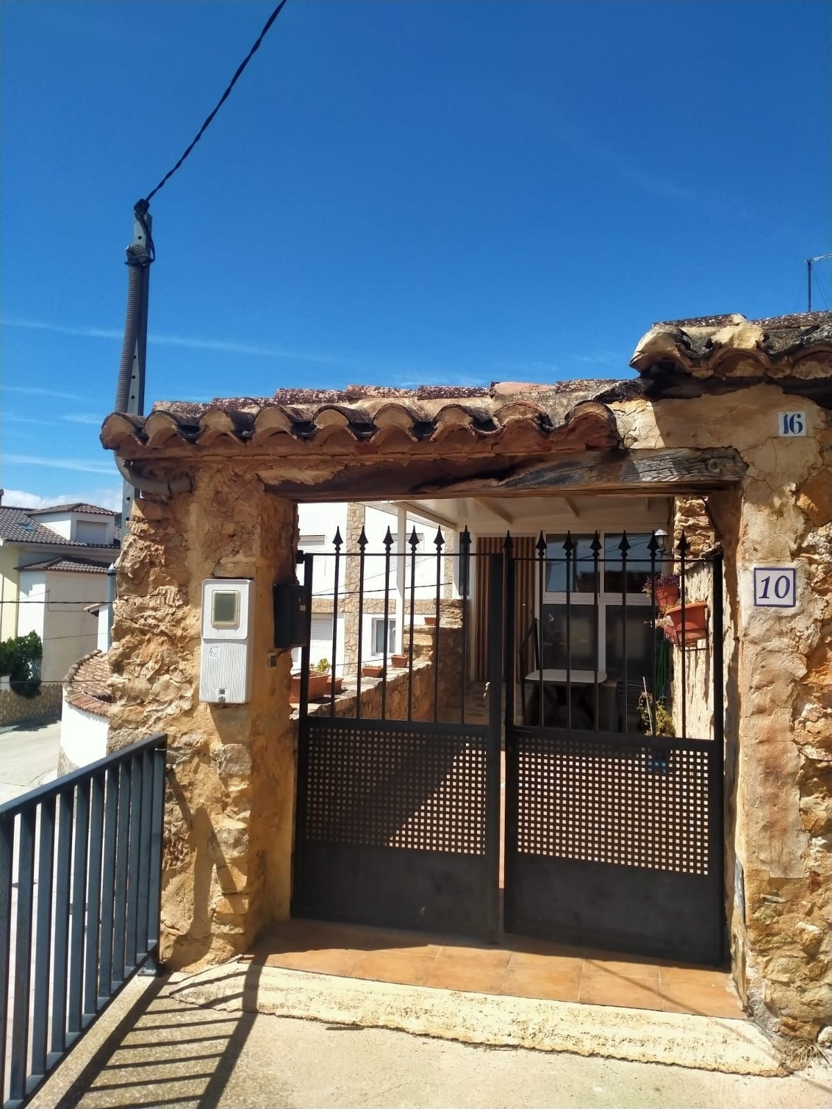The entrance gate features a wrought iron design, leading to a stone facade with a traditional roof. Sunshine casts a bright glow over the exterior, with a glimpse of a path that offers access to the home. The surrounding area consists of residential buildings.