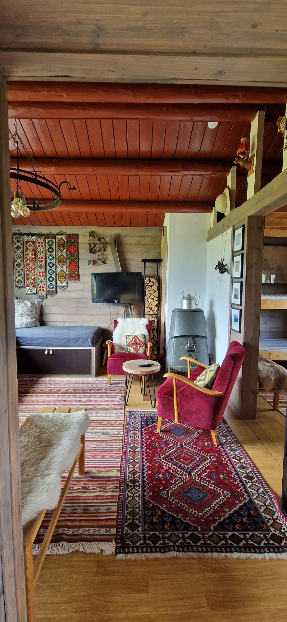 A cozy living area is showcased, featuring a rich area rug and a mix of seating options. A vibrant red armchair and a small table are positioned near a decorative wood-burning stove. Wall art adds character, while a warm wood ceiling enhances the inviting space.