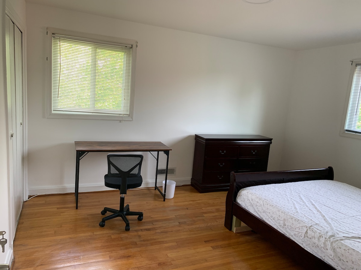 A bright, minimalist bedroom is presented with a spacious bed and a dark wooden dresser. Natural light enters through two blinds-covered windows, illuminating a simple desk and a black office chair positioned near the wall. The warm wooden floor complements the room's decor.