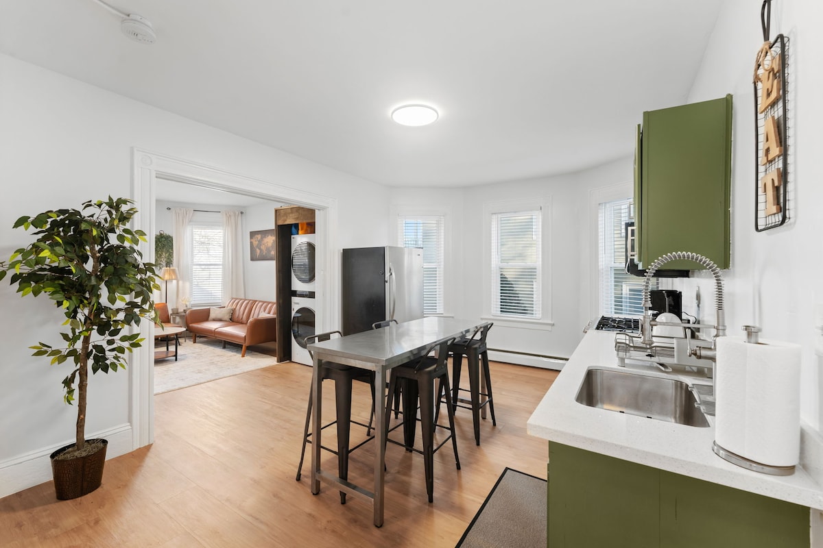 A modern kitchen area is presented with an island featuring four metal stools. A stainless steel refrigerator and stove are visible, along with a sink integrated into a polished countertop. Natural light enters through multiple windows, enhancing the spacious environment.