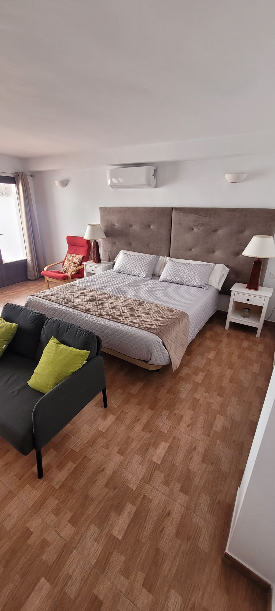 A bright bedroom features a spacious bed with light-colored linens and a textured headboard. A soft throw blanket is draped over the bed. A small seating area includes a gray sofa with green cushions, while a side table stands next to the bed. Natural light enters through glass doors.