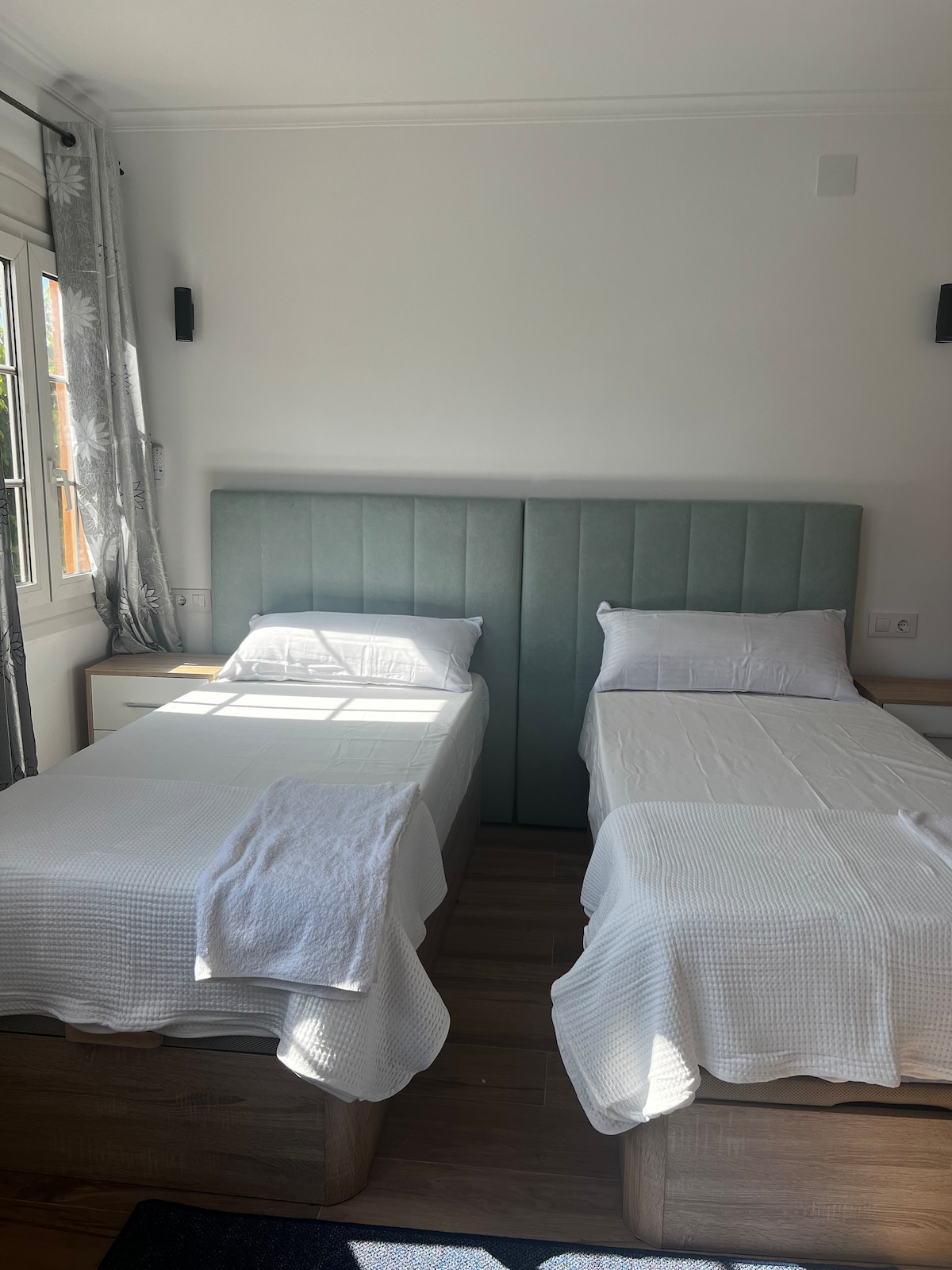 Two individual beds are placed in a well-lit bedroom, featuring light-colored bedding and a soft green headboard. Natural light filters in through the window, illuminating the simple decor and wooden flooring.