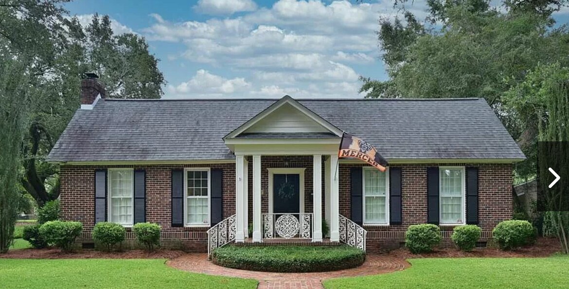 A charming brick house features a well-maintained front yard with neatly trimmed hedges and two symmetrical bushes. A welcoming pathway leads to the entrance, adorned with a decorative railing. Large windows provide natural light, and a flag adds a touch of character to the exterior.