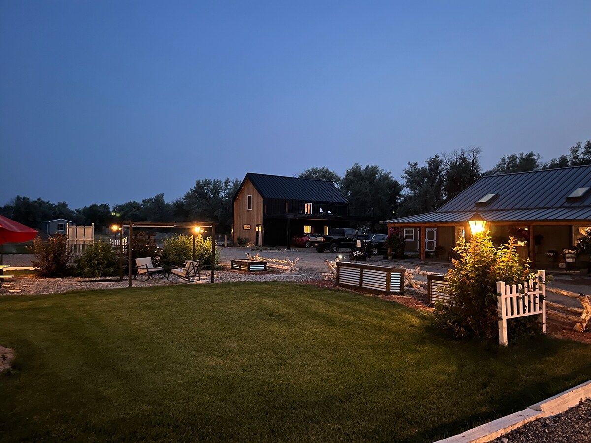 A spacious outdoor area is illuminated by warm lighting, highlighting a well-maintained lawn and pathways. Surrounding structures include a two-story building and a single-story facility, both featuring metal roofs. Seating areas with picnic tables and a fire pit are visible, creating a welcoming communal space.