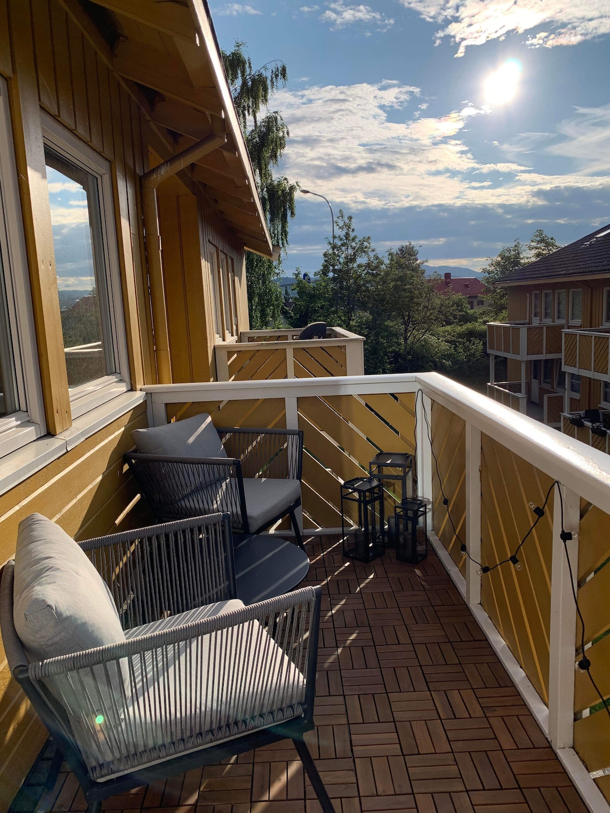 A balcony is shown with two black chairs and a round table, surrounded by a wooden railing painted in yellow. Soft sunlight bathes the area, highlighting the wooden flooring and greenery in the background. Decorative lanterns sit on the floor, enhancing the space.