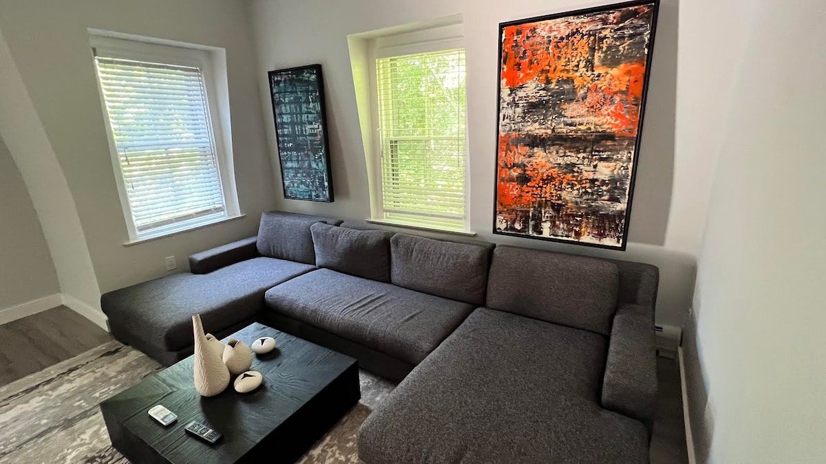 A modern living area is highlighted by a spacious gray sectional sofa arranged around a dark coffee table. Two large artworks adorn the walls, providing a vibrant contrast to the neutral tones. Natural light filters through the window blinds, creating a bright atmosphere.